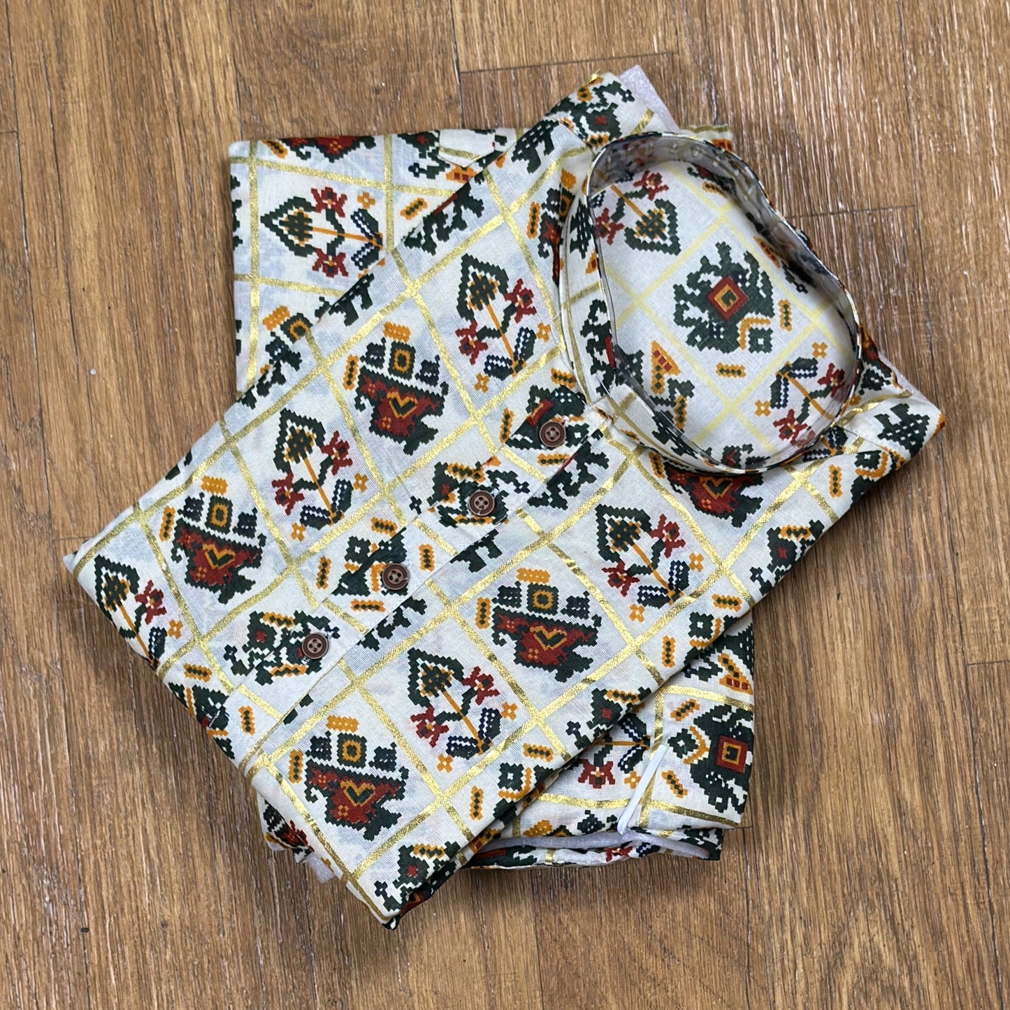 Folded mandarin collar shirt with green, red, and yellow geometric floral print on white fabric, placed on wooden floor